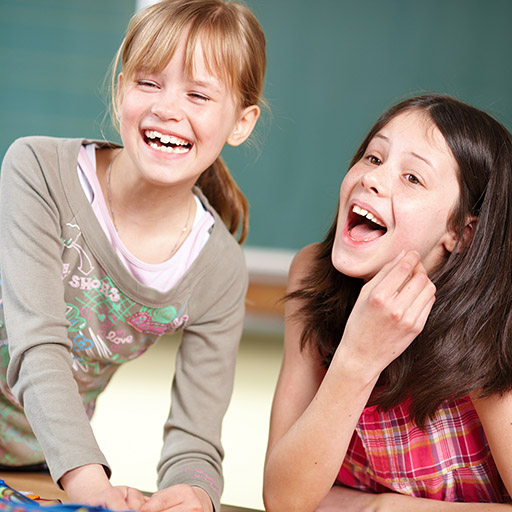 Zwei Mädchen lachen gemeinsam im Klassenzimmer vor einer Tafel.